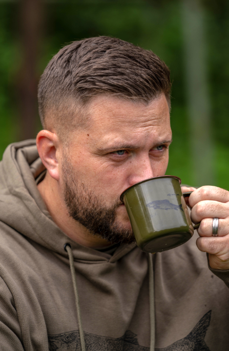 Чаша KORUM Classic Enamel Mug Barbel - 330ml, мъж пие от емайлирана чаша по време на риболов.