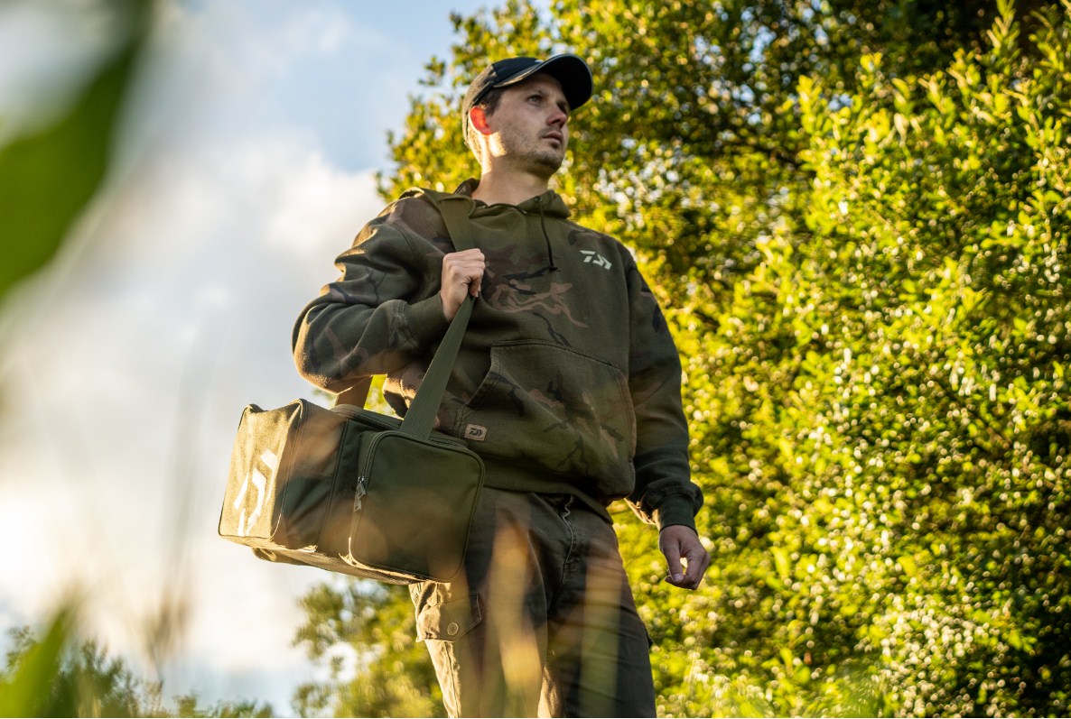 Сак DAIWA BLACK WIDOW COMPACT TACKLE BAG being carried by a man in a natural outdoor setting.