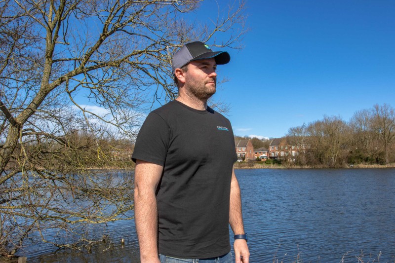 Тениска PRESTON Black T-Shirt - черен цвят - XXXL на мъж край водоем, с акпликирано лого на Preston Innovations.