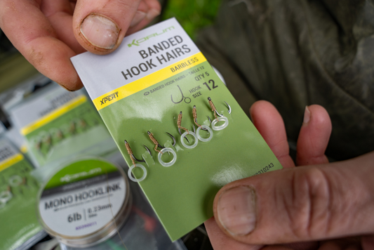 Ръце държат опаковка KORUM Banded Hook Hairs с пет куки и ясно видими прозрачни ластичета за стръв.