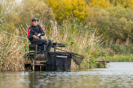 Риболовец на платформа Daiwa D500SB край водата, със стъпенка, странична маса и подредени принадлежности.