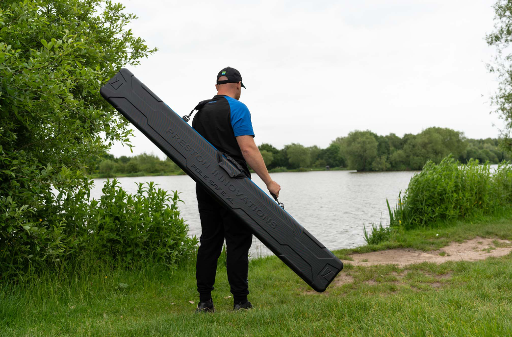 Мъж с твърд калъф за щеки PRESTON Hardcase Pole Safe XL, стоящ до водоем в природата.