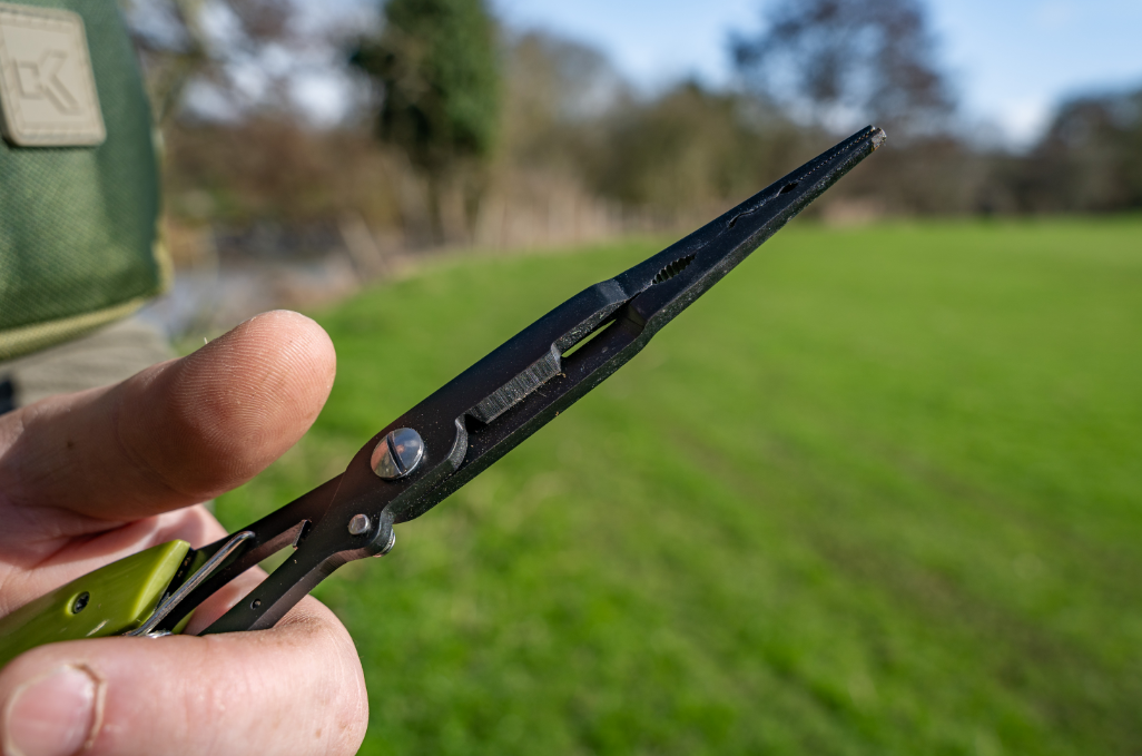 Мултифункционални клещи с карабинер KORUM Multi Tool With QC Lanyard, показани в ръка на фон от зелена трева.