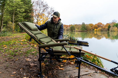 Луксозен фидер стол KORUM S23² Supa Deluxe Accessory Chair на риболовно място с мъж, държаща стол.