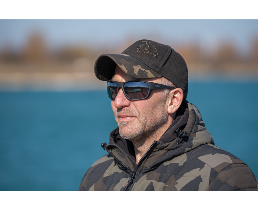 Мъж с поляризирани слънчеви очила AVID CARP SeeThru OptiWrap Grey Lens, стоящ на открито до вода.