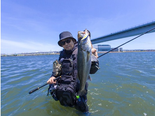 Риболовец с улов на спининг въдици за морски риболов DAIWA LATEO, стоящ във водата под мост.
