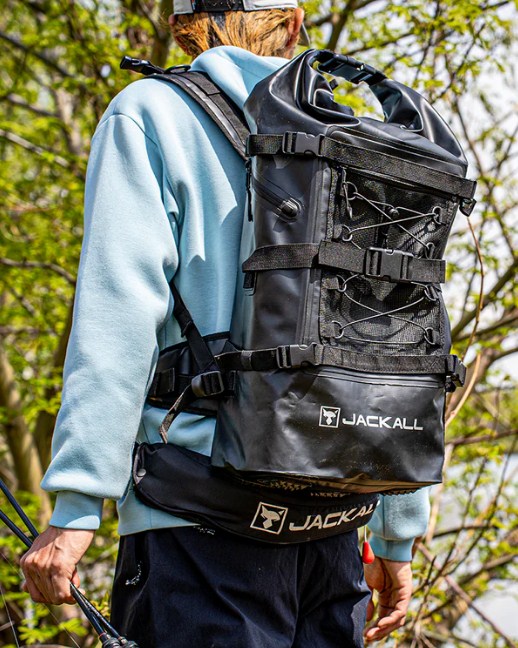 Водоустойчива чанта за спининг риболов JACKALL WATERPROOF BACKPACK L (30L - 35L) - Черна на човек, носеща риболовна екипировка.