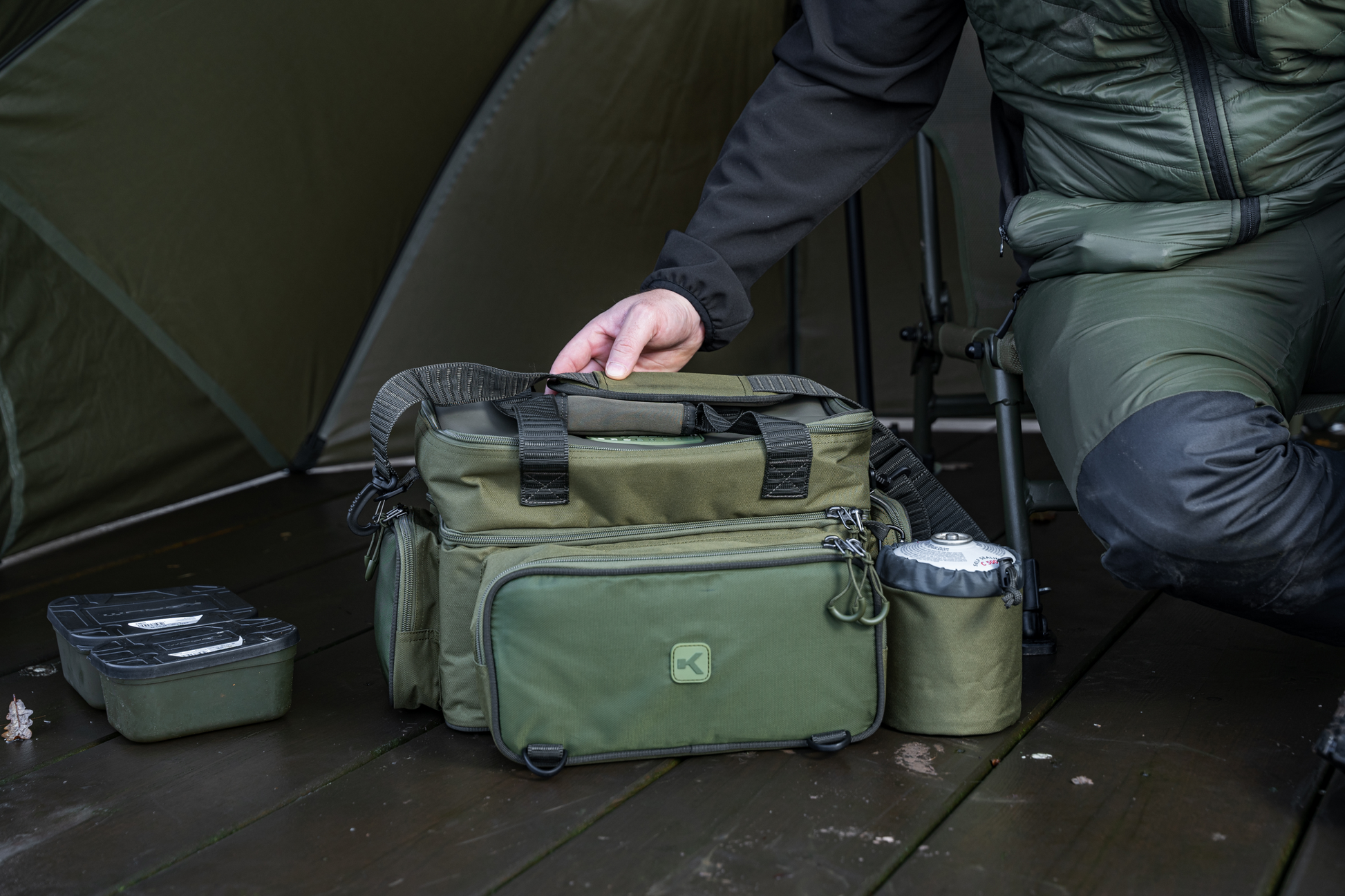 Чанта KORUM Progress Tackle & Bait Bag на дървена настилка, носена от човек в палатка.