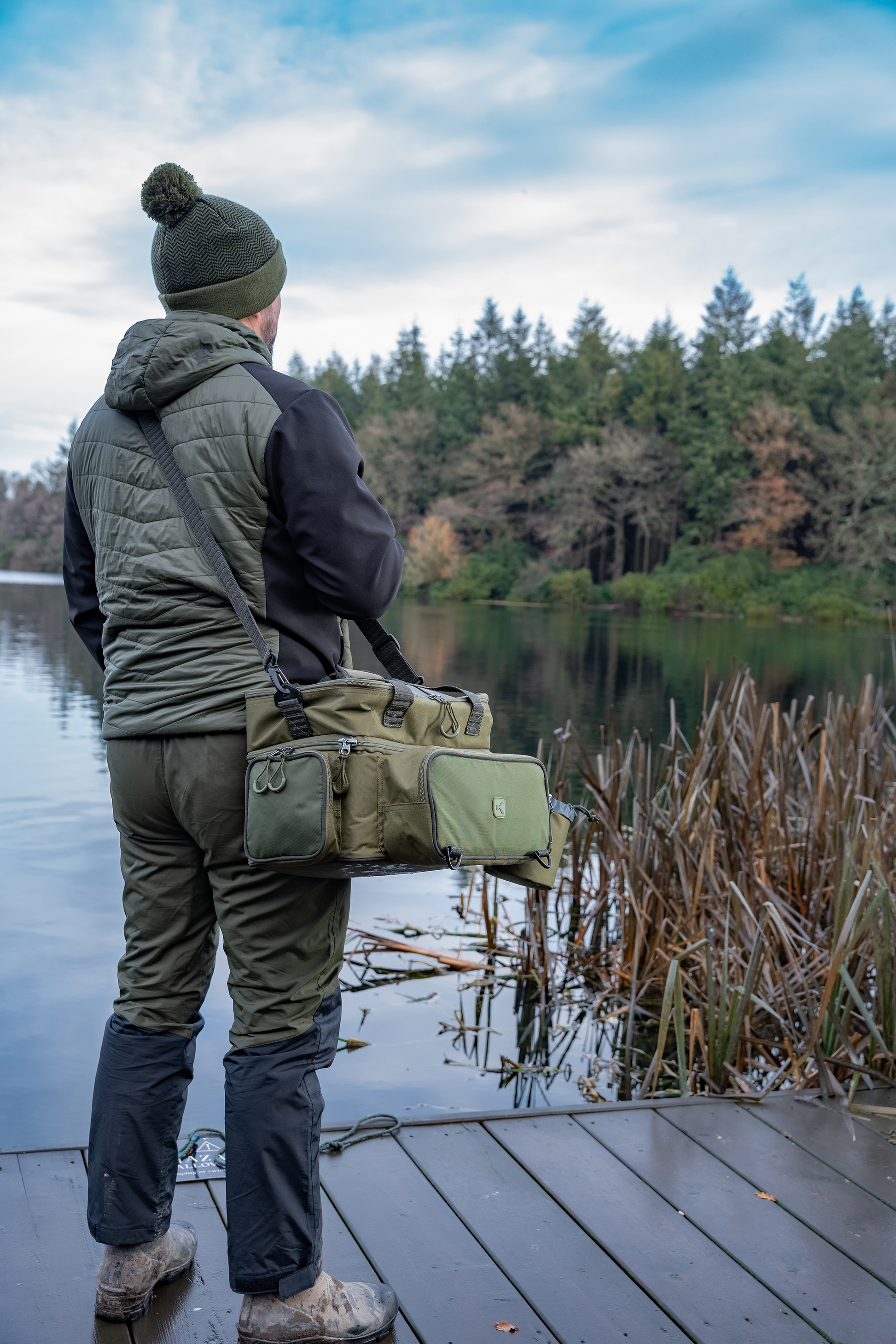 Мъж с чанта KORUM Progress Tackle & Bait Bag, спохождаща на езеро със зеленина около него.