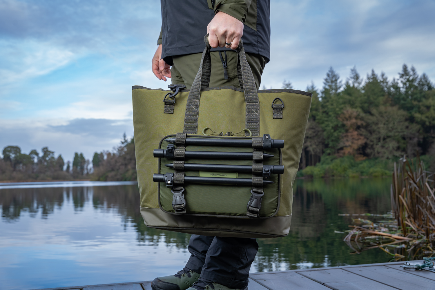 Чанта KORUM Progress Bait & Bits Bag - 25L held by a person near the water in a scenic outdoor setting.