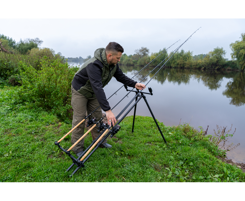 Родпод Korum DELUXE 3 ROD TRIPOD, човек настройва рибарска стойка на брега на река.