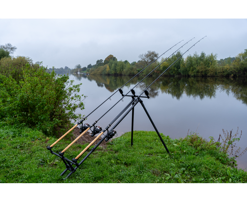 Родпод Korum DELUXE 3 ROD TRIPOD с три въдици на ръба на река в зелен пейзаж.