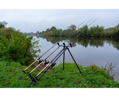 Родпод Korum DELUXE 3 ROD TRIPOD с три въдици на ръба на река в зелен пейзаж.