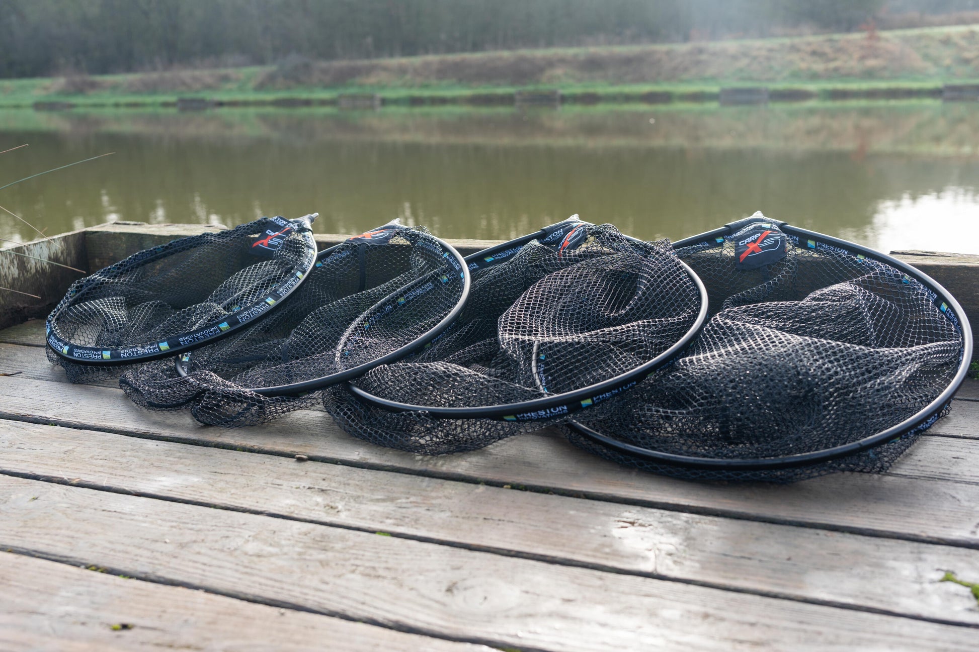 Глава за кеп PRESTON Carp XS Landing Net displayed on a wooden dock by a tranquil water surface.
