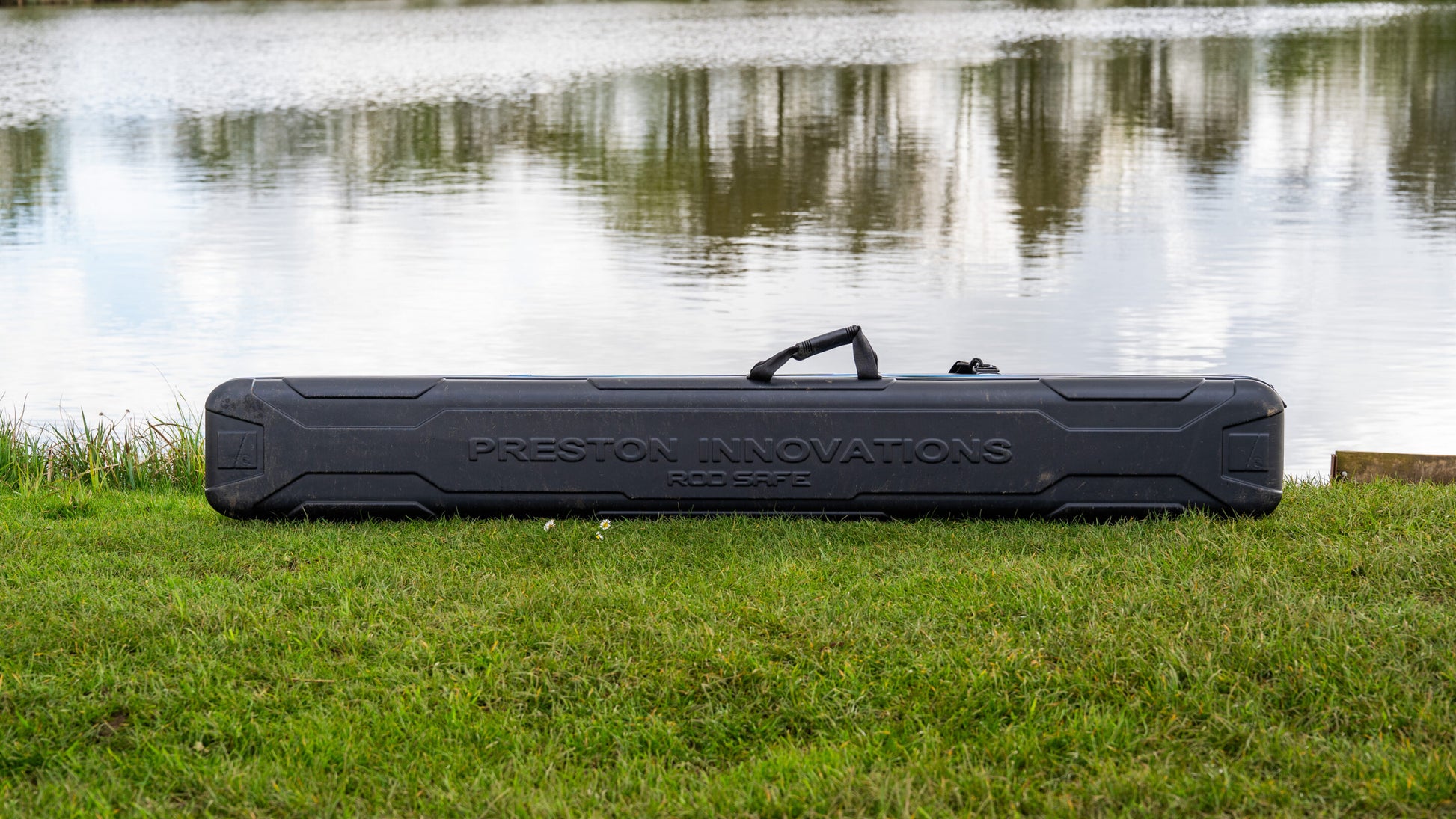 Твърд калъф PRESTON HARDCASE ROD SAFE лежи на трева до водоем, осигуряващ защита за въдици.