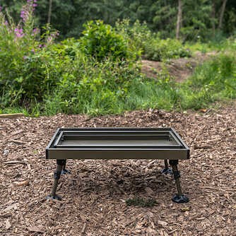 Двуетажна маса AVID Double Decker Bivvy Table in a natural outdoor setting on wood chips.