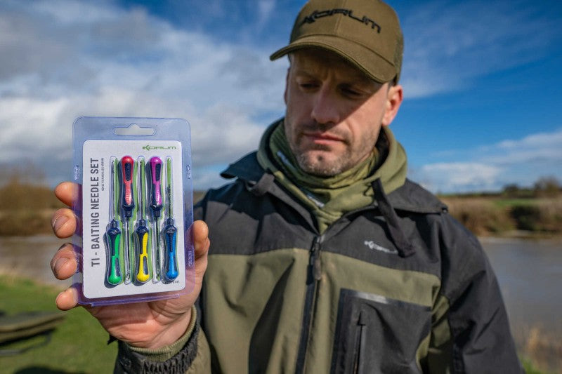 Мъж държи комплект игли KORUM TI - BAITING NEEDLE SET на фона на река и синьо небе.